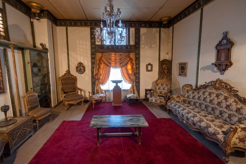 An interior salon with period furniture, upholstered seating, chandeliers, display cabinets, and a red carpet inside the Gayer-Anderson Museum, Cairo