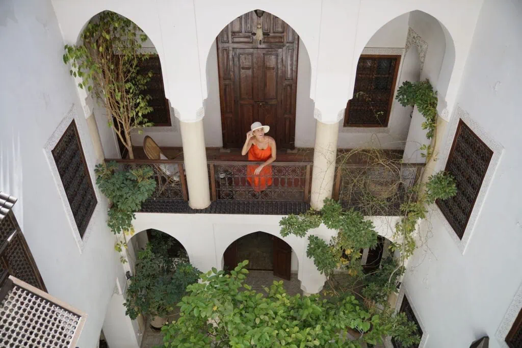 Riad courtyard balcony with a woman standing by carved wooden railings overlooking a tiled courtyard with plants and a central fountain, Marrakech
