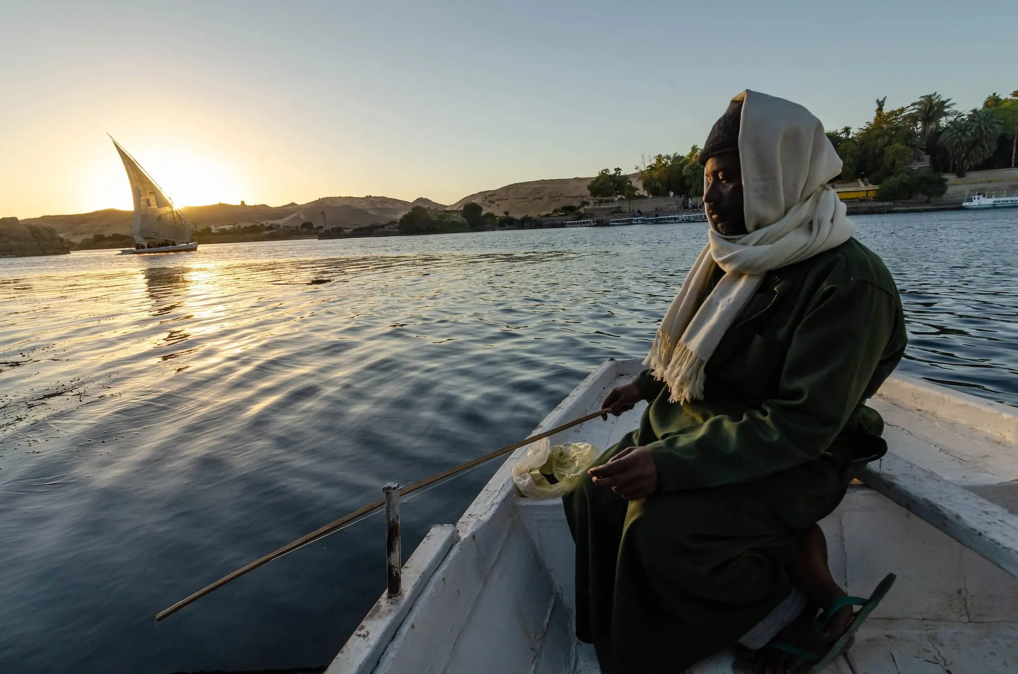 Nubian fisherman in traditional white headwrap sitting in boat on Nile River at sunset with felucca sailboat in background