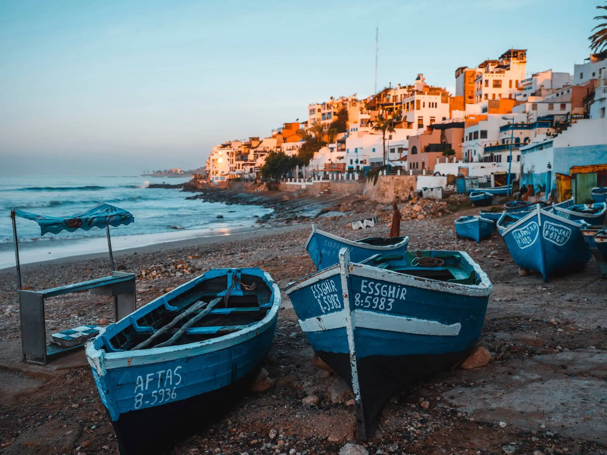 Traditional blue fishing boats on beach in Taghazout village with white buildings on cliffs