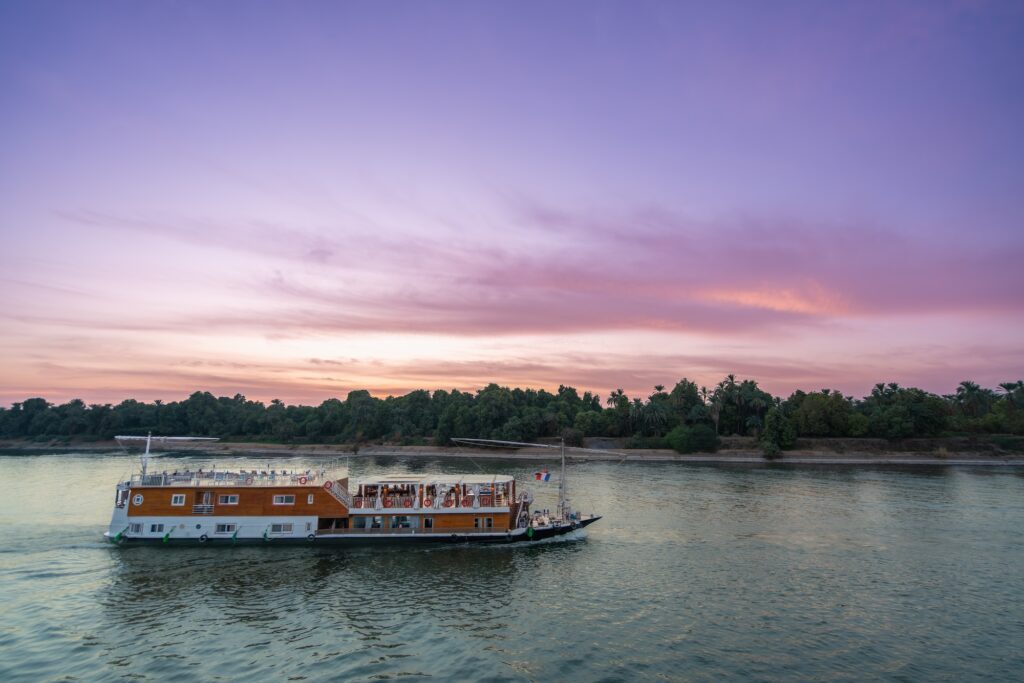 A traditional dahabiya sailboat gliding on calm river waters as warm sunset light reflects across the surface, Nile River, Aswan
