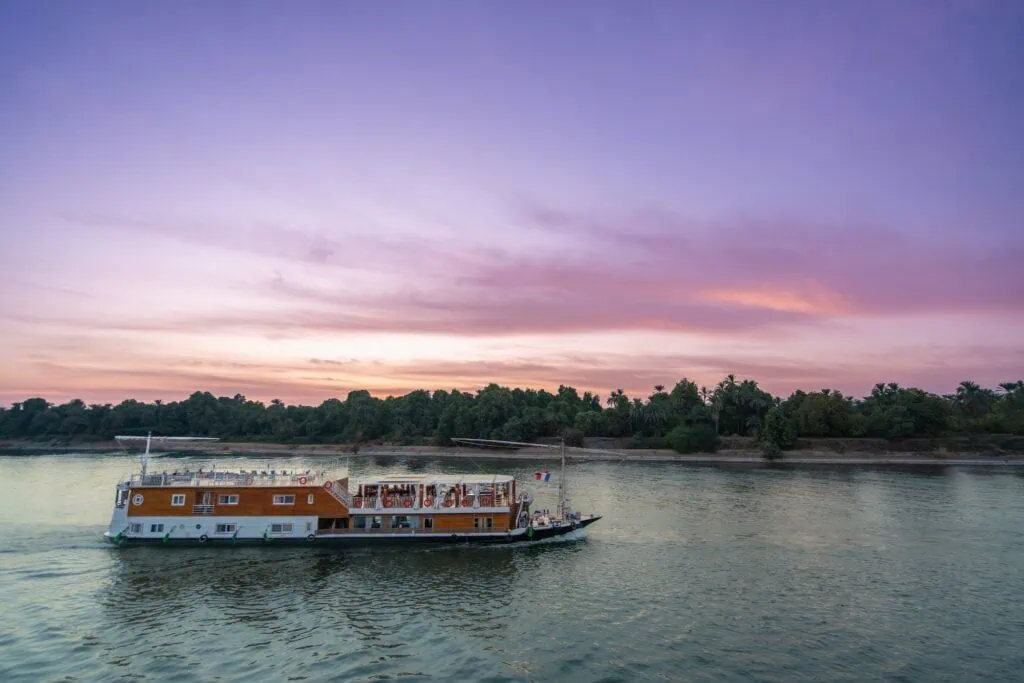 A traditional dahabiya sailboat gliding on calm river waters as warm sunset light reflects across the surface, Nile River, Aswan