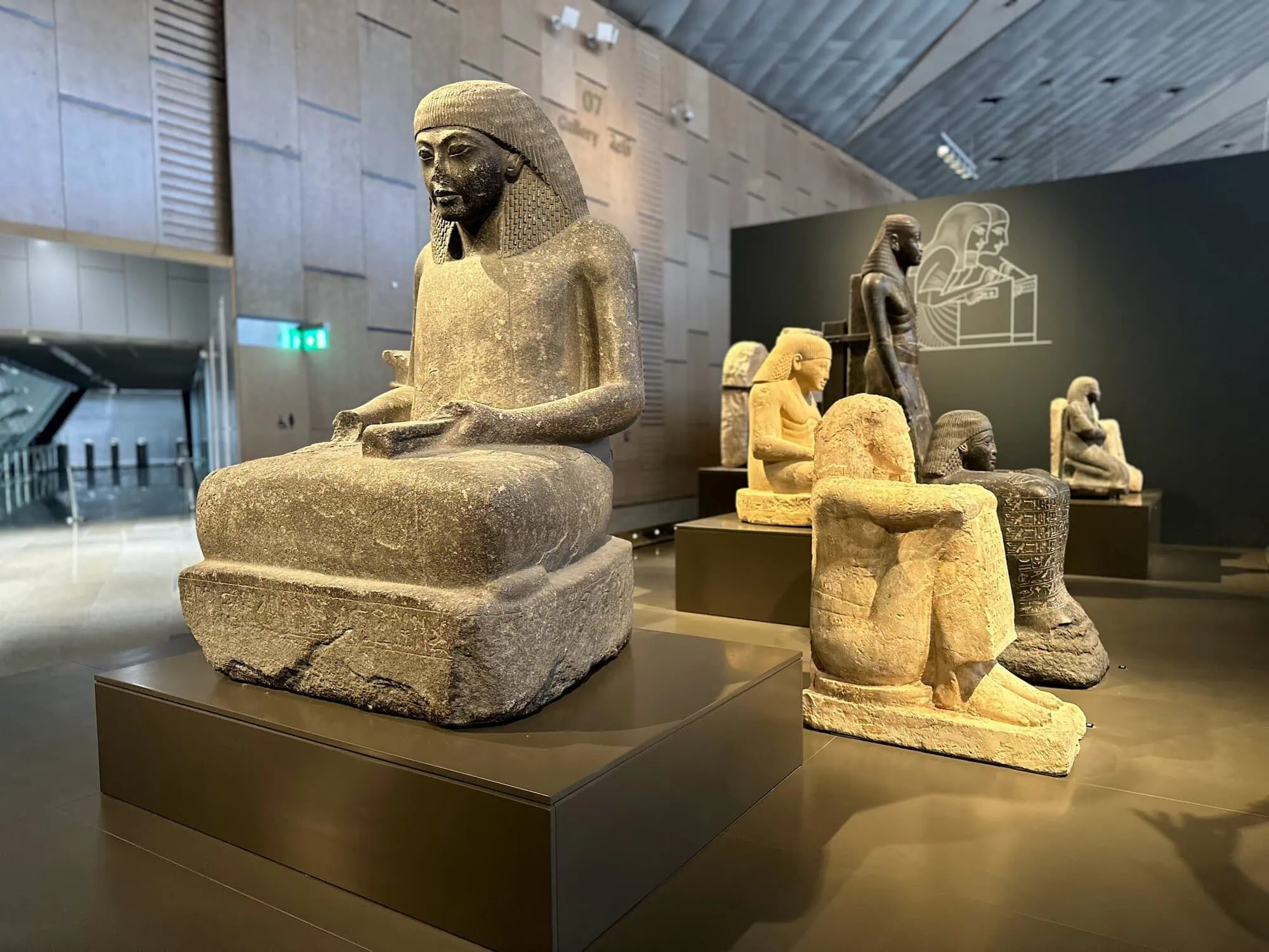 Interior gallery view inside the Grand Egyptian Museum showing ancient Egyptian statues, carved artifacts, and stone fragments displayed on pedestals throughout a modern museum hall.