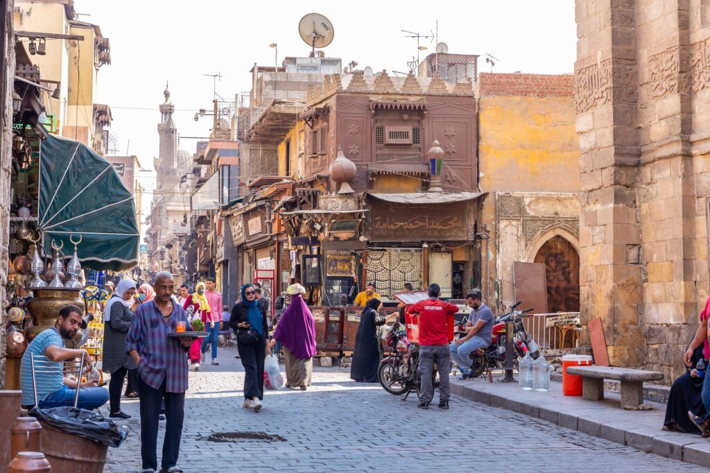 Khan el-Khalili bazaar with craft and souvenir shops, pedestrians, surrounding historic buildings, and Al-Azhar Mosque in the background, Cairo