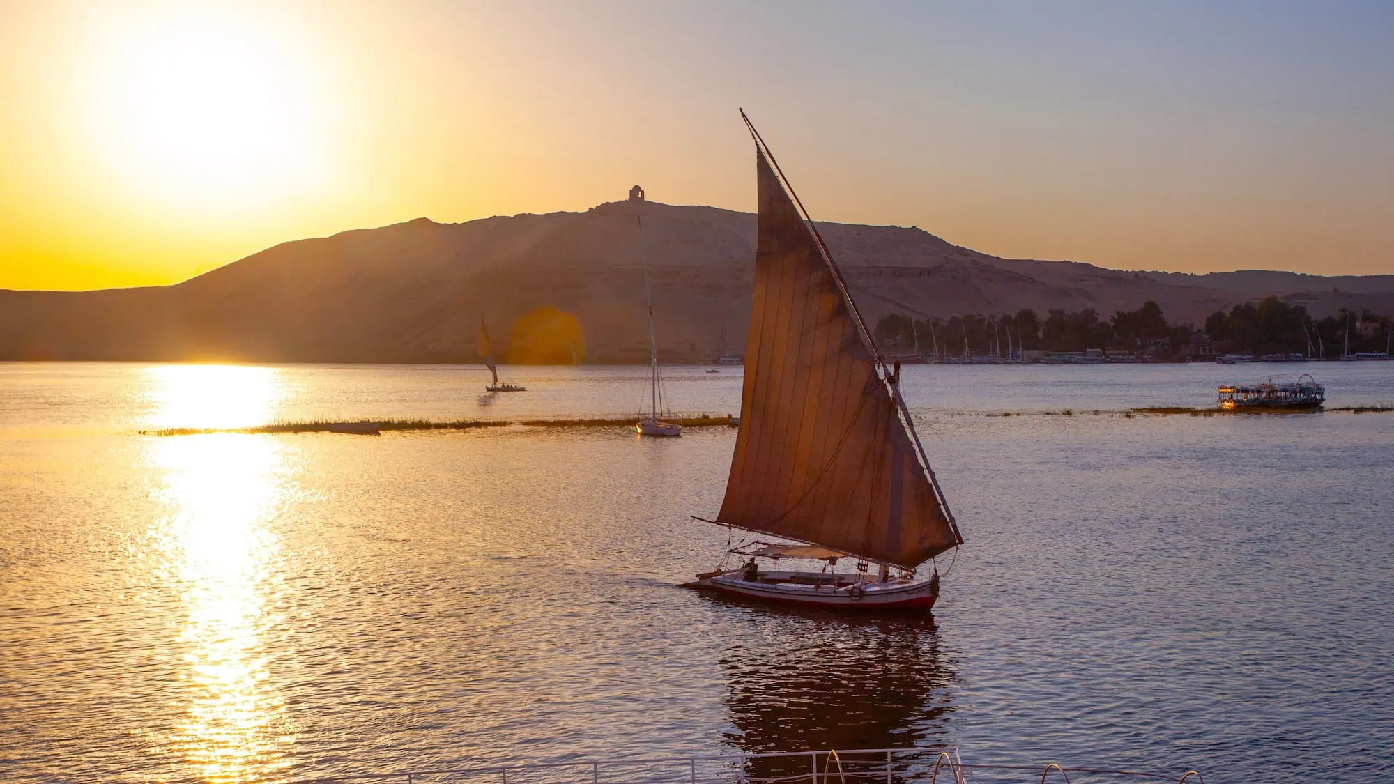 Traditional felucca sailboat on the Nile River at sunset with mountains in background