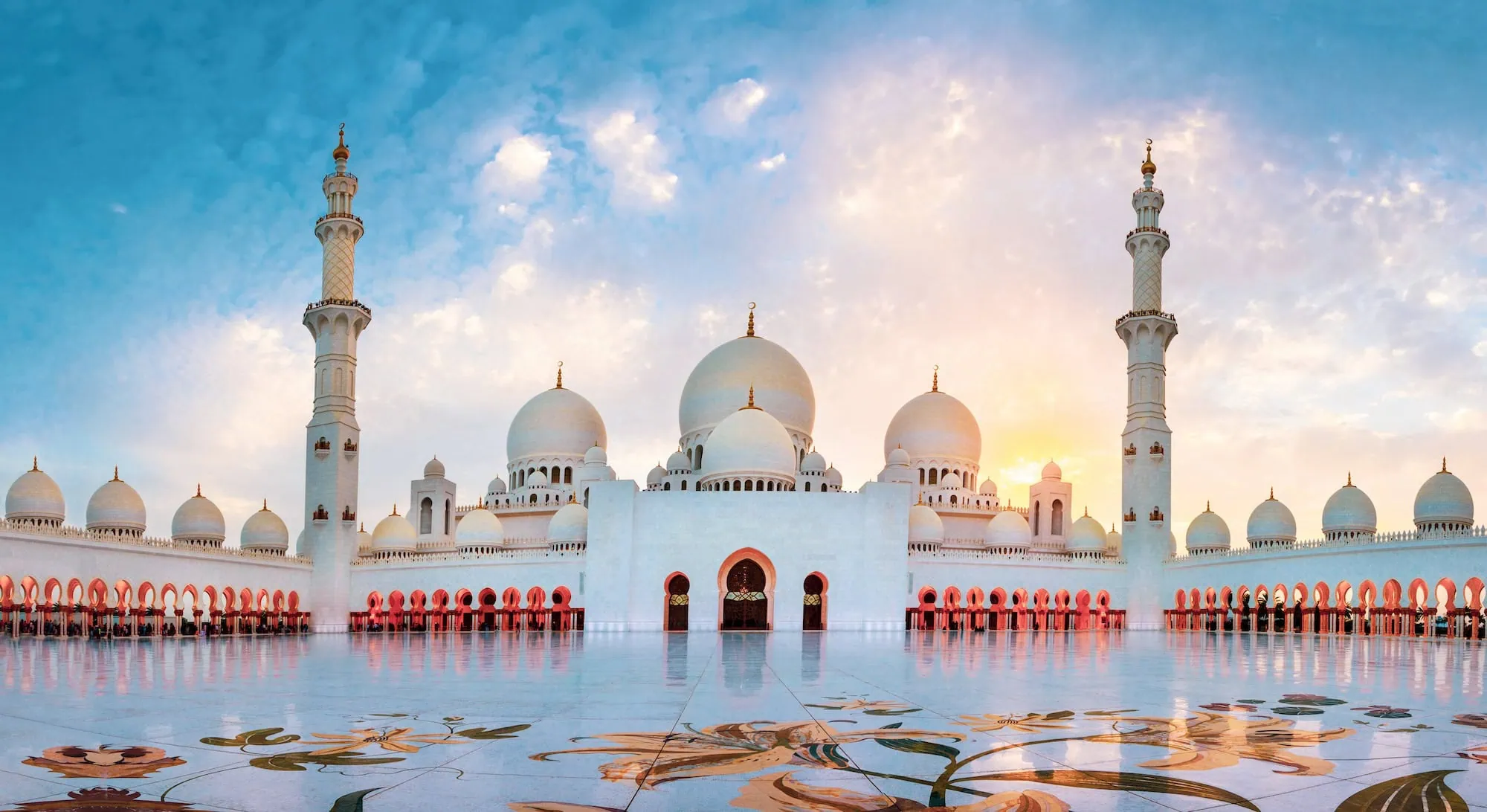 Panoramic sunset view of Sheikh Zayed Grand Mosque with white domes and minarets reflected in courtyard pool