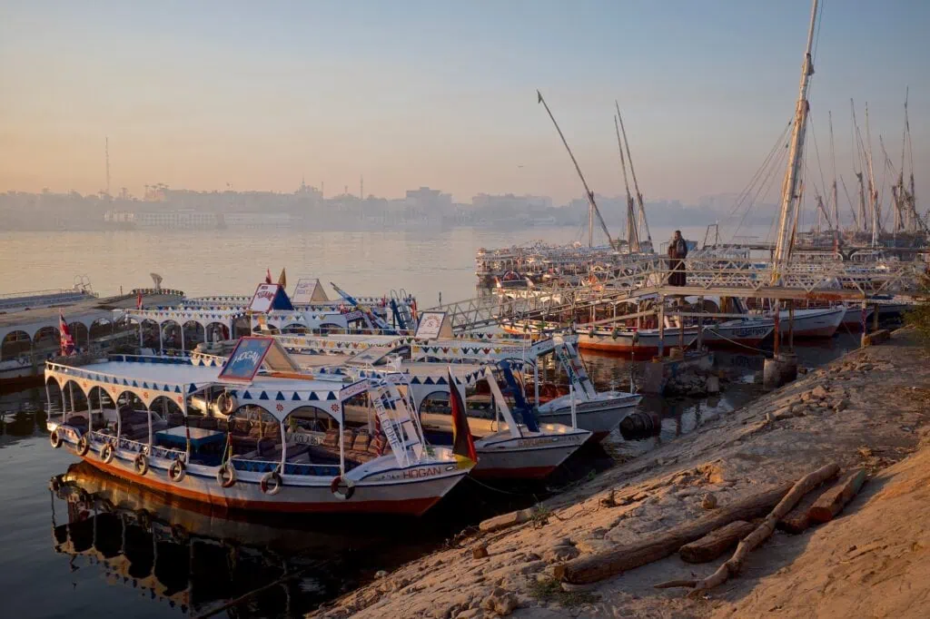 Tourist boats docked along the west bank of the Nile River with mooring posts and riverfront view, Luxor