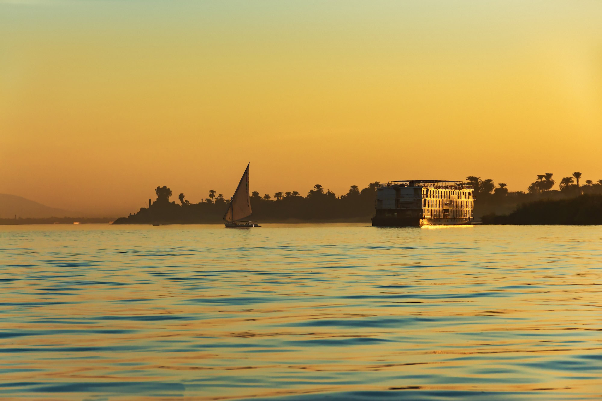 Traditional sailboat and cruise ship on the Nile River at sunset in Luxor, Egypt with golden sky reflection