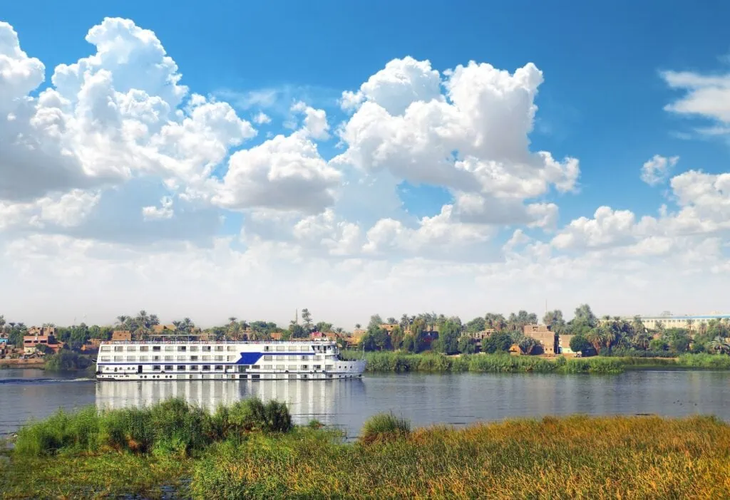 A Luxury Nile Cruise vessel on the Nile seen from the river bank, with the far bank behind it, the five-star floating hotel used on the classic Egypt tour route.