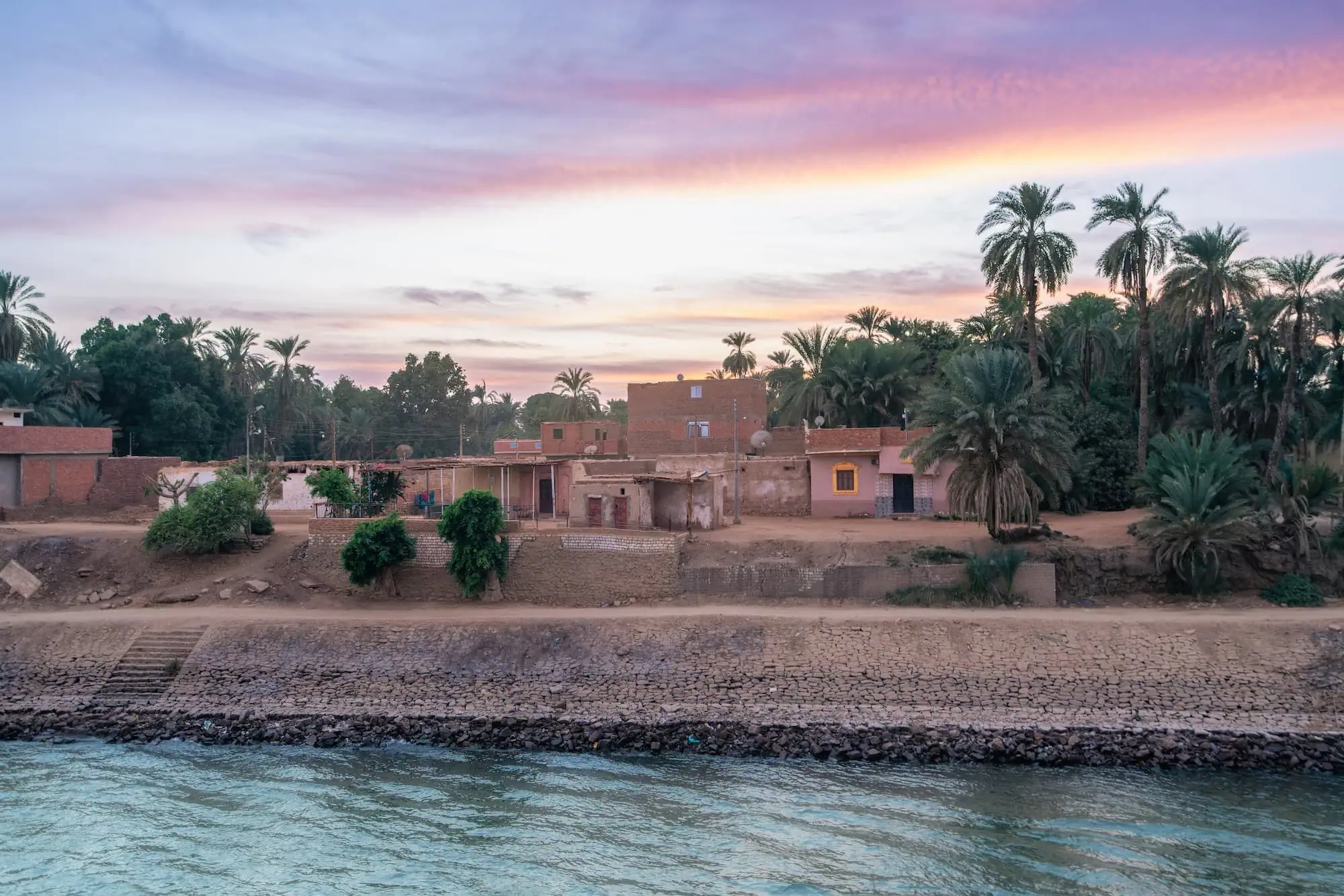 Traditional Egyptian village along the Nile River at sunset with palm trees and ancient buildings