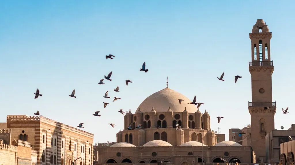 A flock of pigeons flying past the minarets and courtyard of Al-Azhar Mosque near Khan El Khalili, Cairo