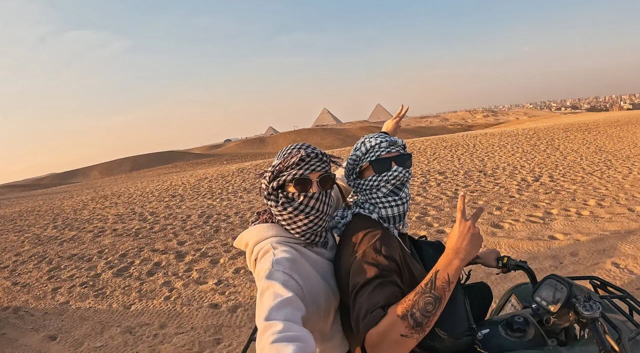 Couple taking selfie while riding quad bike and making peace sign with giza pyramids in background during sunset