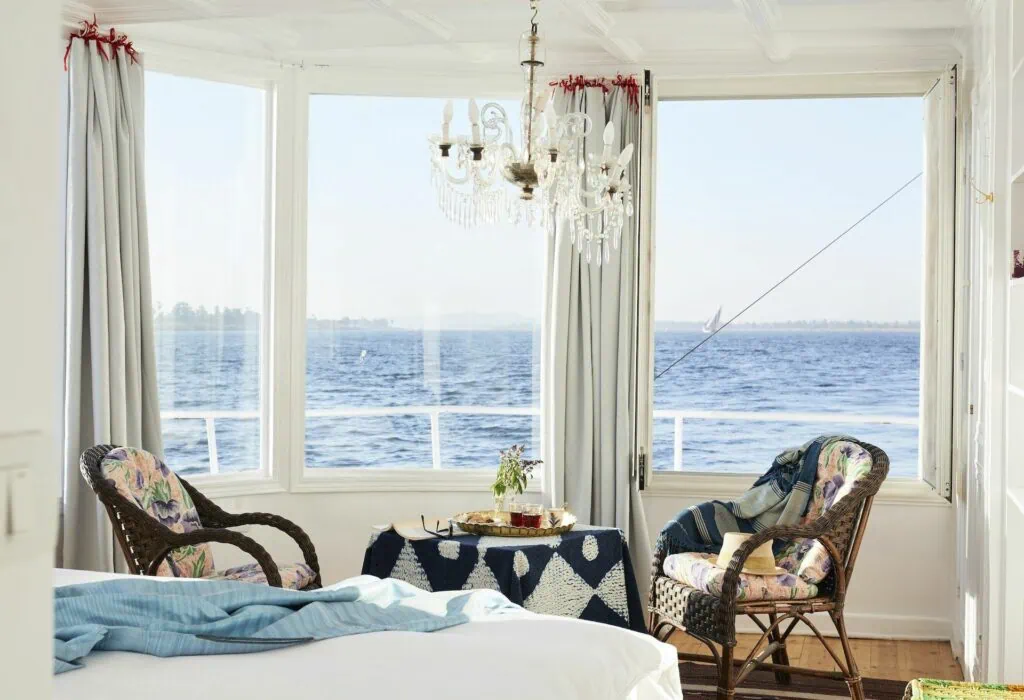 Nour El Nil Dahabiya suite interior with double bed, wooden furnishings, seating area, and river-view windows, Aswan