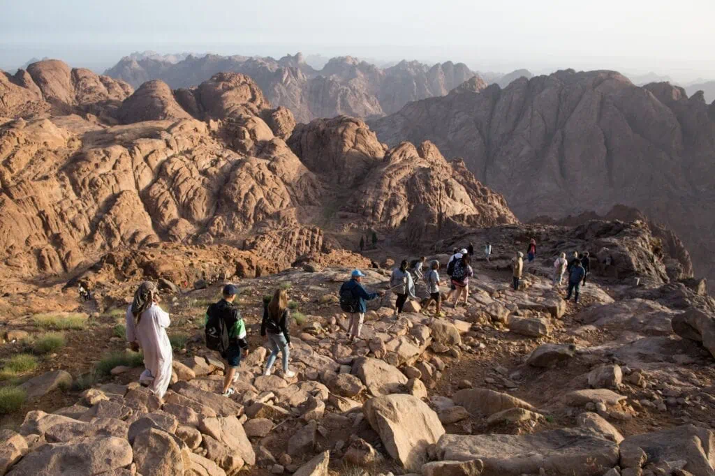 Tourists walking along a rocky trail at sunrise with surrounding granite peaks on Mount Sinai, Saint Catherine