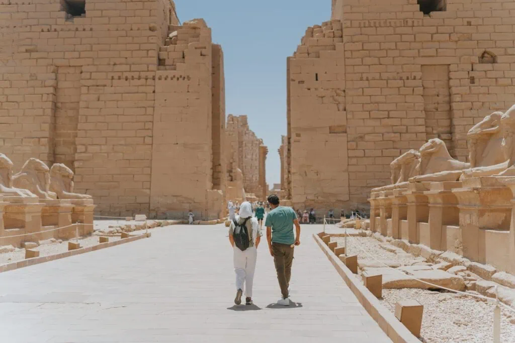 Karnak Temple with tourists walking between large sandstone walls and ram-headed sphinx statues, Luxor