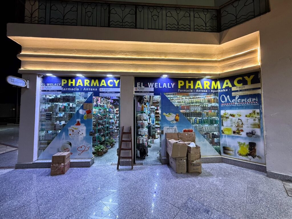Exterior of El Welaly Pharmacy illuminated at night with visible stocked shelves inside, Sharm El Sheikh