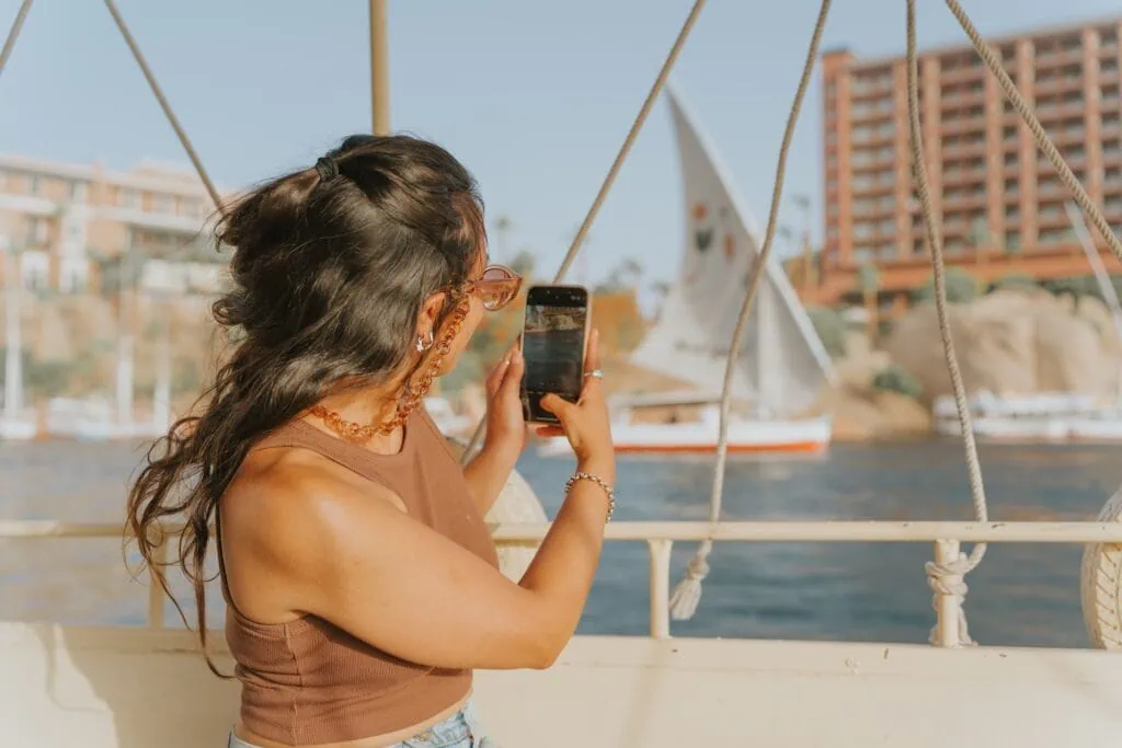 Traditional felucca sailboat on the Nile River with a woman photographing the scene on a smartphone, Aswan