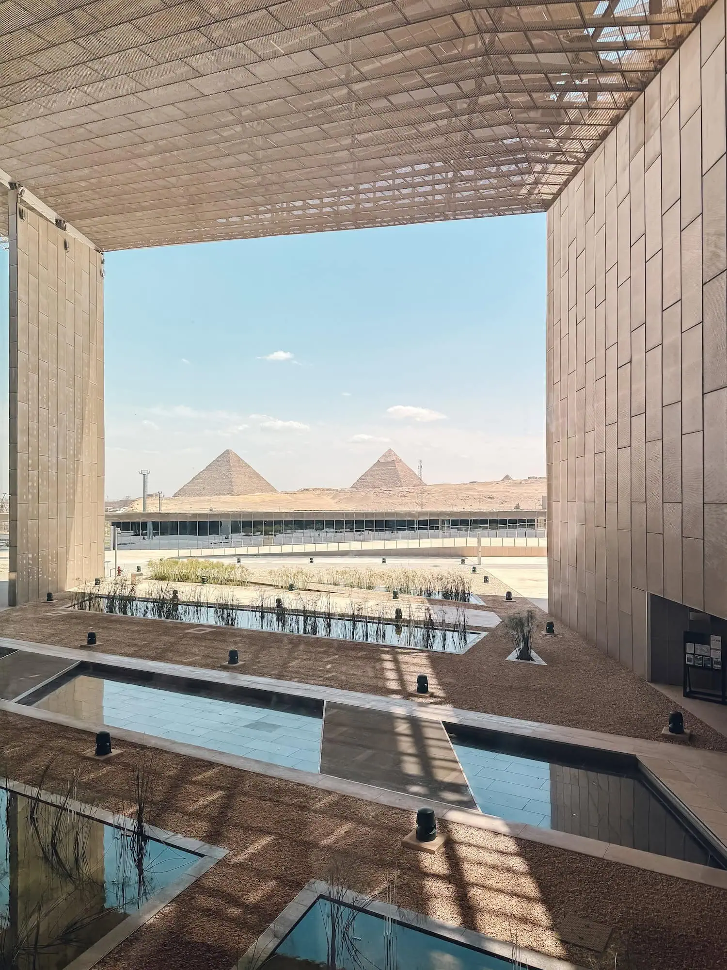 Interior view of the Grand Egyptian Museum's main atrium with the three Pyramids of Giza framed through the architectural window, the fully-opened museum at the foot of the Giza plateau holding the complete Tutankhamun collection of 5,000+ artefacts.
