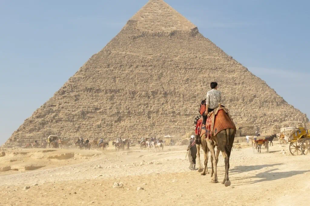 Tourists riding camels near the Great Pyramid of Giza with sandy plateau and clear daytime sky, Giza