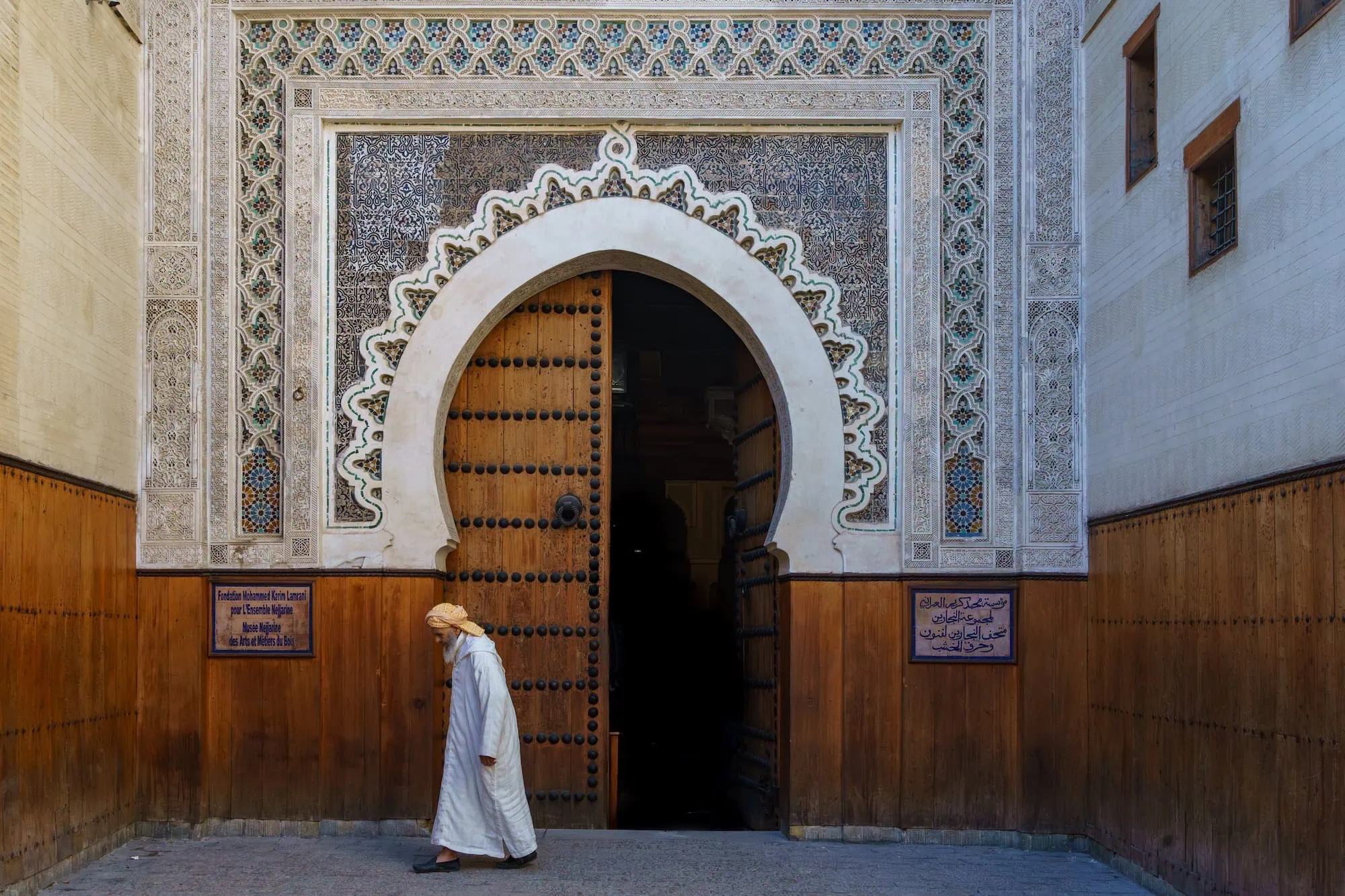 Traditional man in white robes walking past ornate entrance of Nejjarine Museum of Wooden Arts and Crafts in Fez