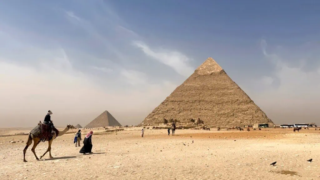 The Great Pyramids of Giza with visitors riding camels across the desert sand in the foreground, on the Giza plateau outside Cairo.