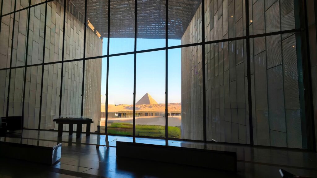 The Great Pyramid framed by the large window and modern interior structure of the Grand Egyptian Museum, Giza