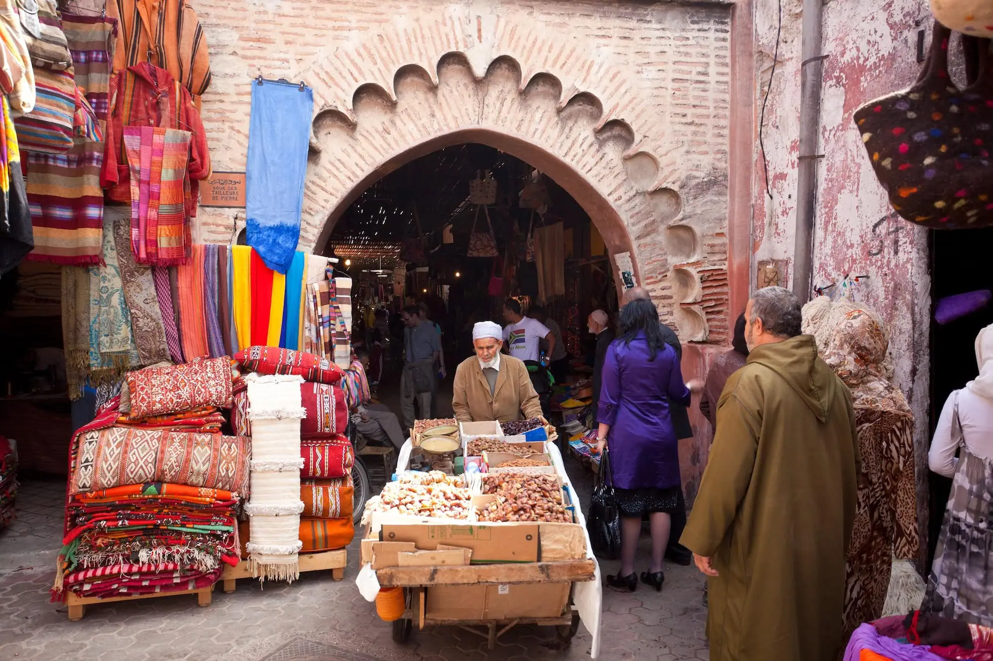 Bustling traditional souk marketplace in Marrakech with vendors selling colorful textiles and shoppers navigating the market
