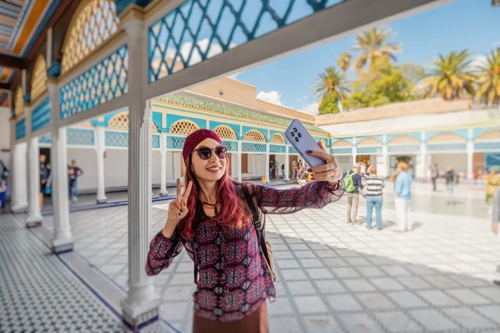 Person taking a selfie with victory sign in the courtyard with tiled floors and arches at Bahia Palace, Marrakesh